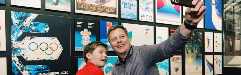 A father and son take a selfies with a Olympic Museum display in the background.